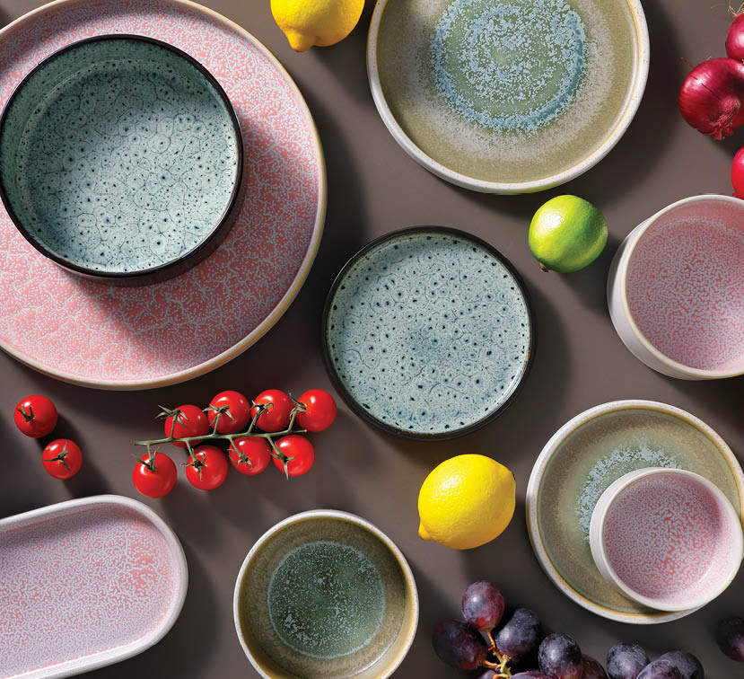 Various ceramic plates and bowls with textured patterns are arranged on a surface alongside lemons, limes, cherry tomatoes, and grapes. Description generated by AI