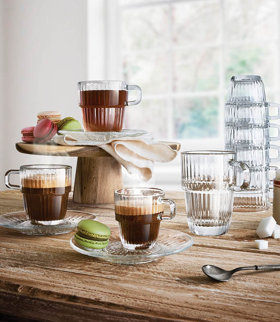 A rustic wooden table holds several clear glass cups filled with coffee, a green macaron on a glass saucer, a stack of upside-down glasses, and a wooden cake stand with a folded cloth and more macarons near a bright window. Description generated by AI