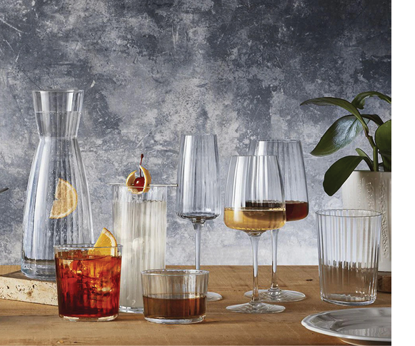 A wooden table holds various clear glassware including a carafe with lemon slices, a tall glass with a cherry garnish, two wine glasses with amber liquid, and other glasses with different beverages, set against a textured gray background with a plant on the side. Description generated by AI