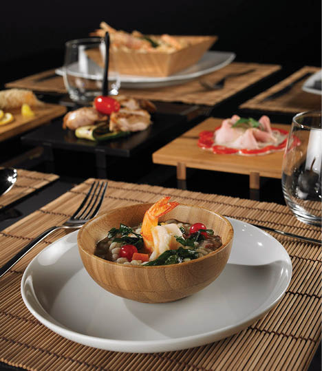 A wooden bowl with shrimp and vegetables sits on a white plate with a fork, knife, and spoon arranged on a bamboo placemat next to a glass of water. Description generated by AI