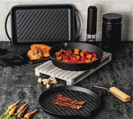 A kitchen countertop displays a cast iron skillet with cooked vegetables, a grill pan, a pepper grinder, a coffee grinder, a halved pumpkin, and some garlic cloves. Description generated by AI