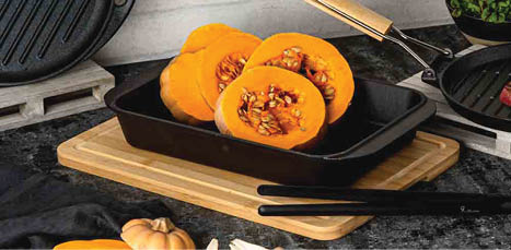 A black roasting pan with halved pumpkins inside sits on a wooden cutting board next to whole and sliced pumpkins on a dark countertop. Description generated by AI