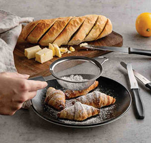 A hand is dusting powdered sugar onto four croissants on a black plate with a metal sieve, with sliced bread and butter on a wooden cutting board in the background. Description generated by AI