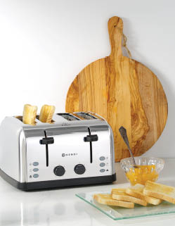 A stainless steel four-slot toaster with two pieces of bread popping up is placed on a white countertop next to a wooden cutting board, a glass bowl of jam with a spoon, and a glass plate holding several slices of bread. Description generated by AI